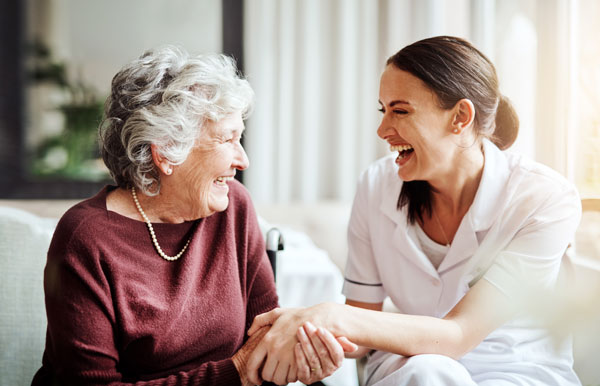 An older lady and a young nurse laughing together while holding hands and sitting.