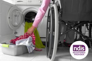A person in a wheelchair putting their laundry in a front-loading washing machine with an NDIS Registered Provider logo showing in the bottom corner