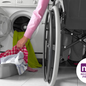 A person in a wheelchair putting their laundry in a front-loading washing machine with an NDIS Registered Provider logo showing in the bottom corner