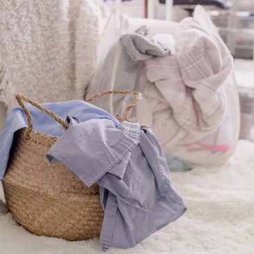 Two baskets filled with laundry. The basket in the foreground is a wicker basket with blue clothes. The basket in the background is a see-through mesh basket with light colour clothes in it to be wash.